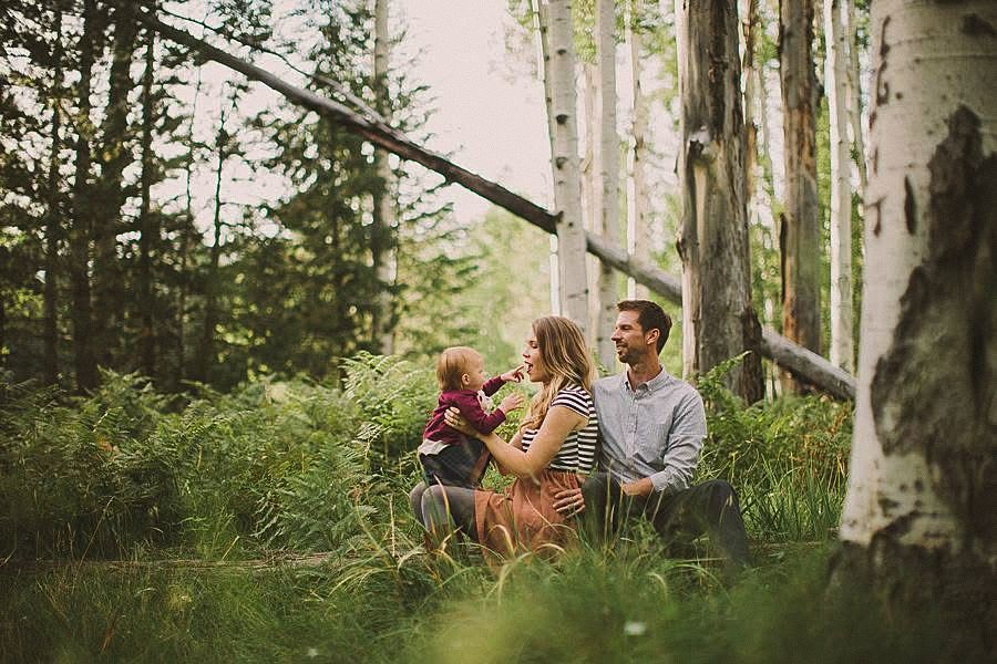 Family of three sitting together in a lush green forest with birch trees, parents holding their baby outdoors.