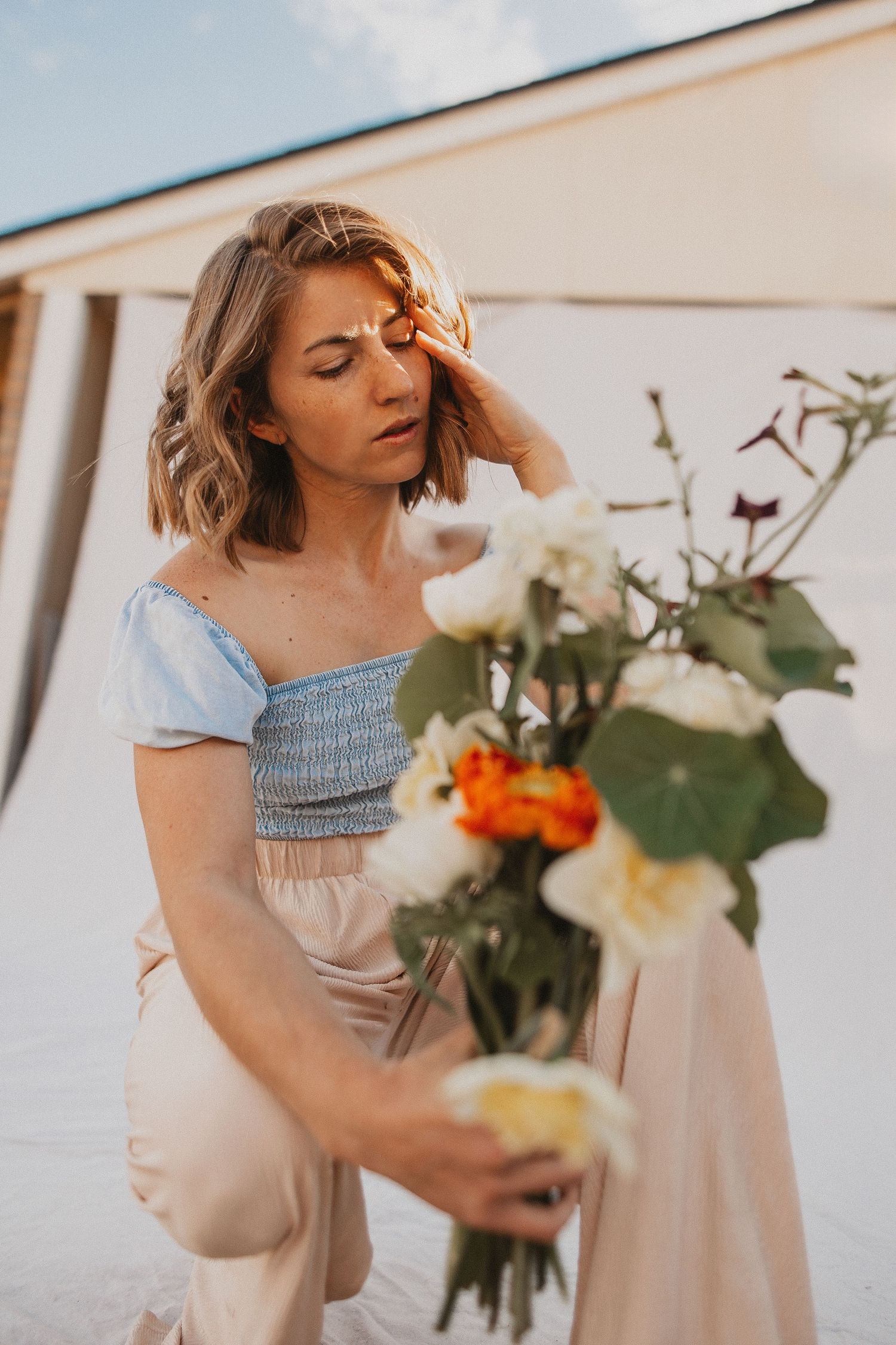 Someone wearing a light colored dress arranges flowers and greenery into a bouquet against a white wall.