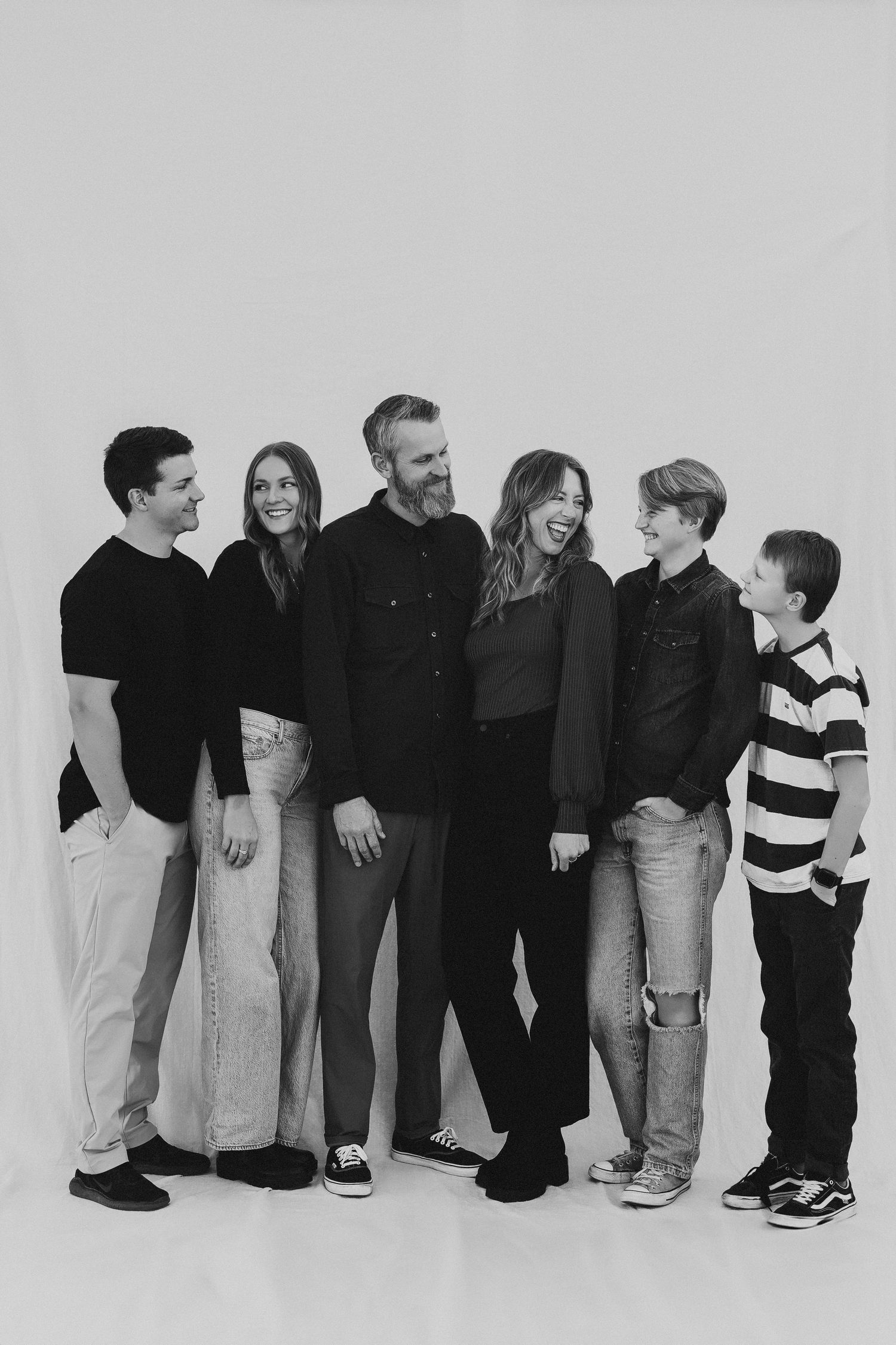 Black and white family portrait of six people smiling and laughing together against a white studio background.