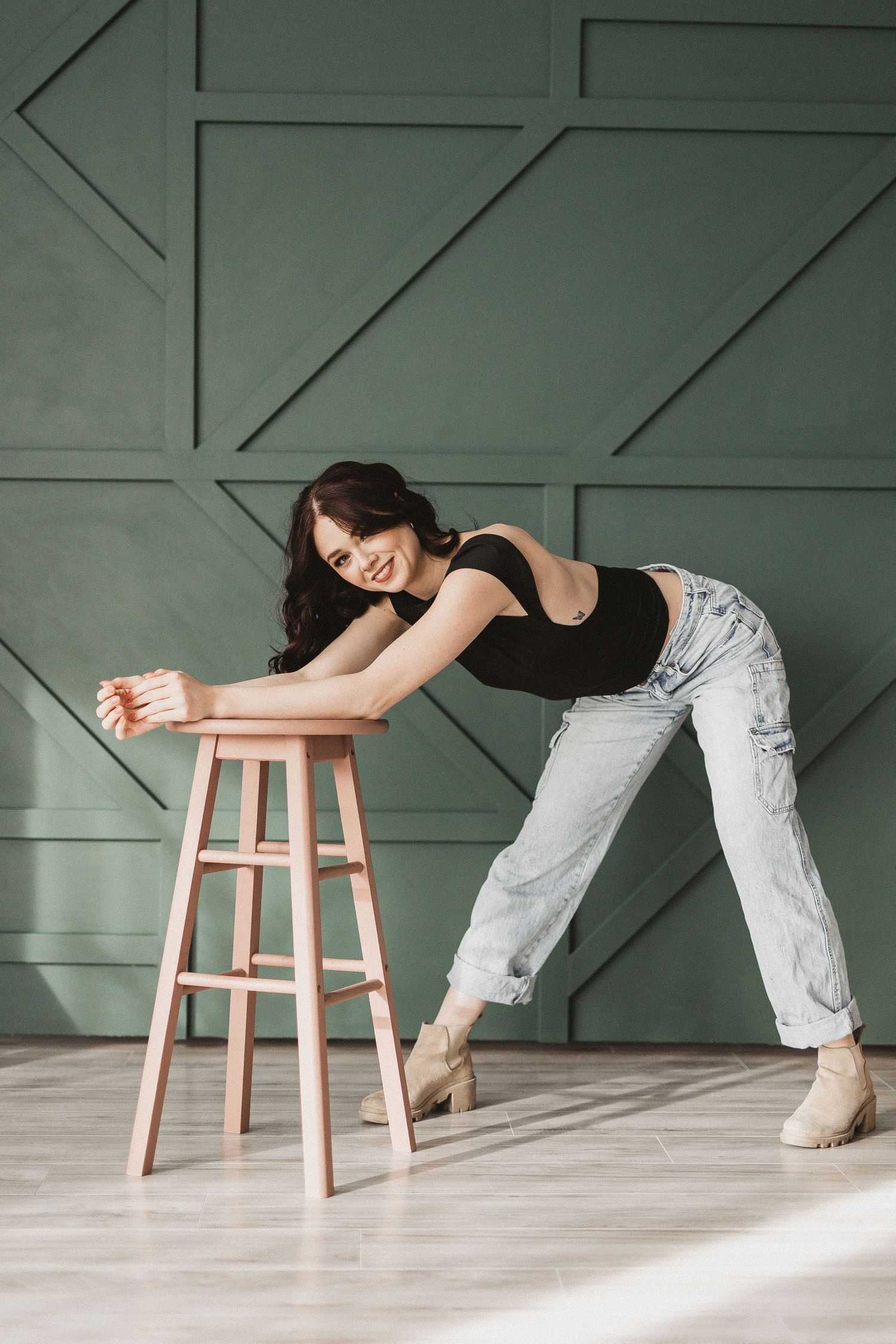 Dancer stretches over wooden stool in dance studio with geometric green wall panels.