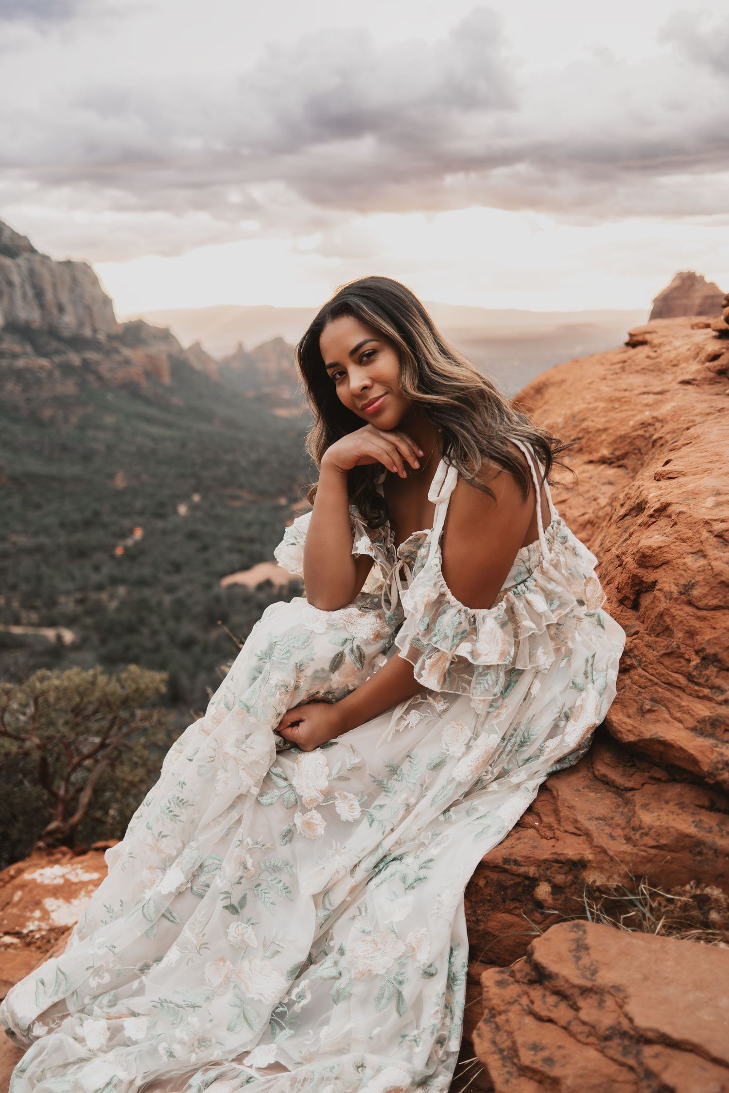 A romantic portrait of someone wearing a flowing white floral dress while sitting on red rocks at sunset in the desert.