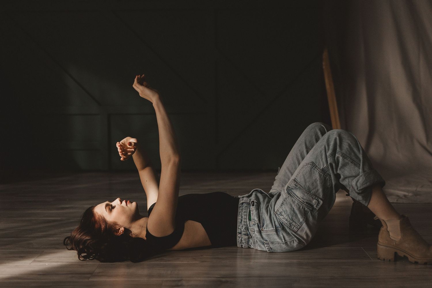 A dancer in a black top and grey skirts performs an expressive floor movement in a dramatic studio setting.