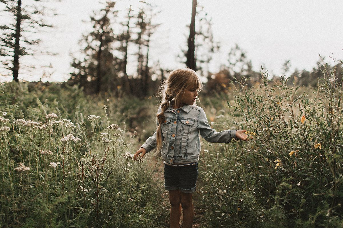 A young girl in a denim jacket walks through a wildflower meadow path surrounded by tall grasses and trees.