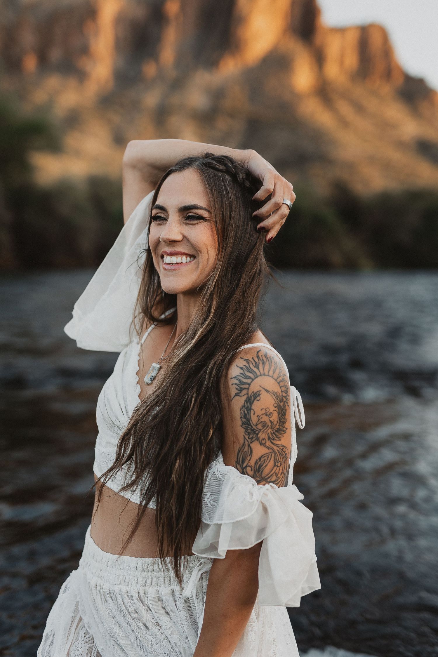 A joyful woman in a white dress with long dark hair and arm tattoo laughs by a riverside at sunset with cliffs behind her.