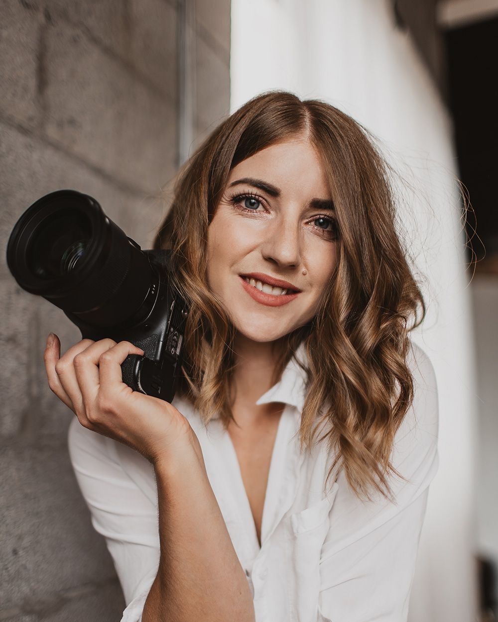 A photographer in a white button-up shirt holds a professional DSLR camera while smiling against a neutral background.