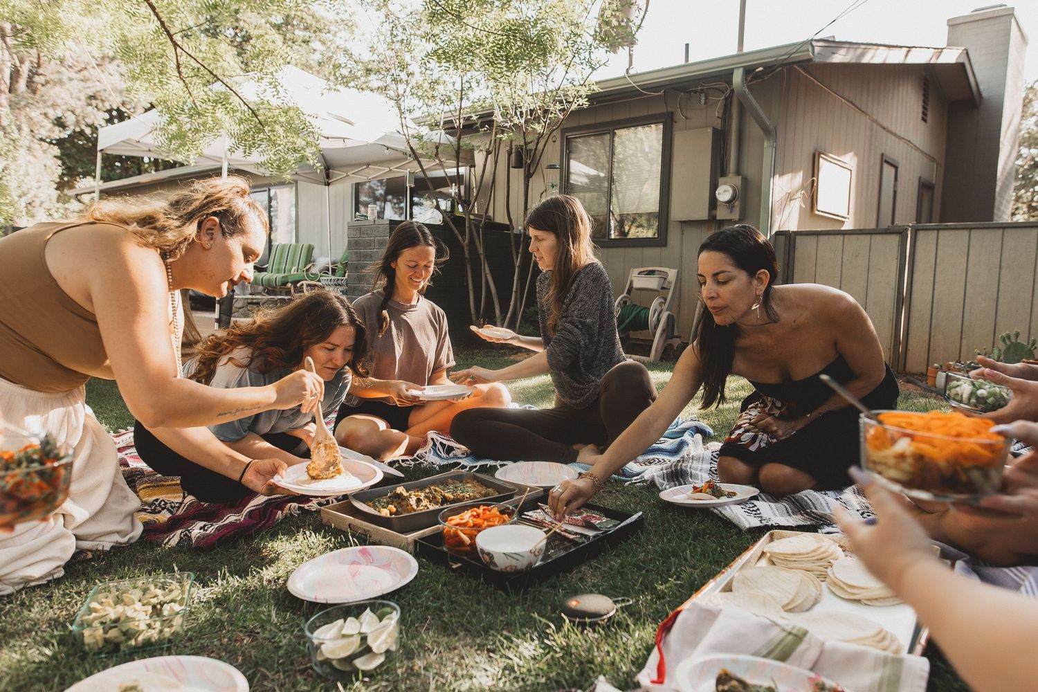 Friends enjoying an outdoor picnic gathering in a backyard with food spread out on blankets during a sunny day.
