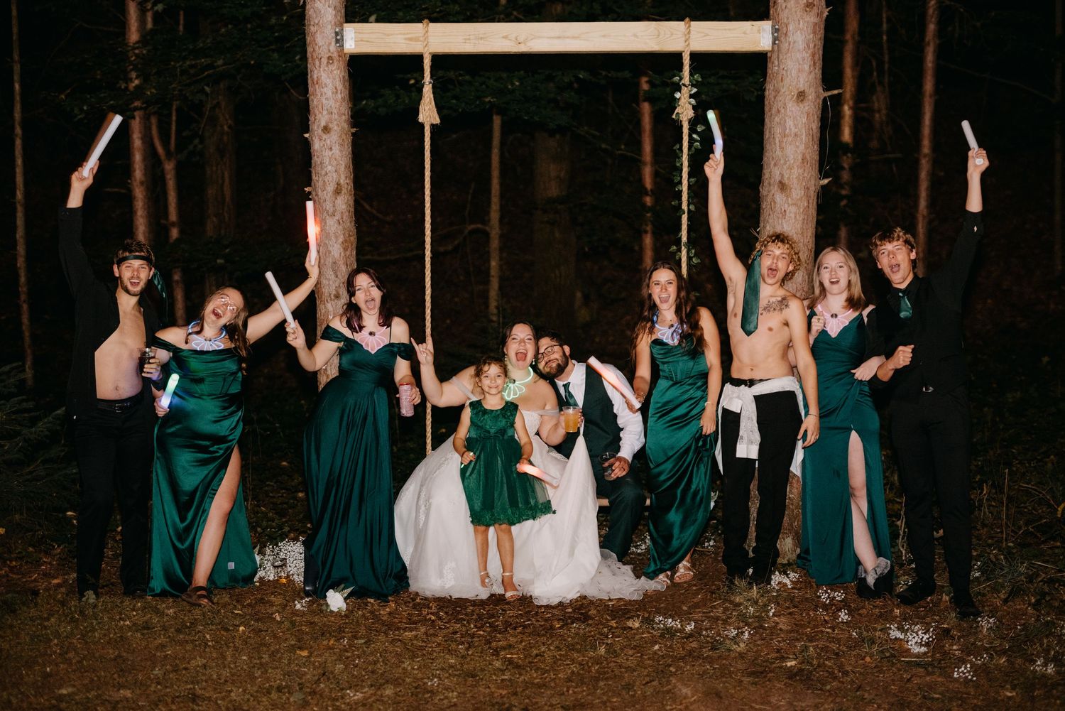 Wedding party in emerald green dresses posing together on swings at night in a forest setting.