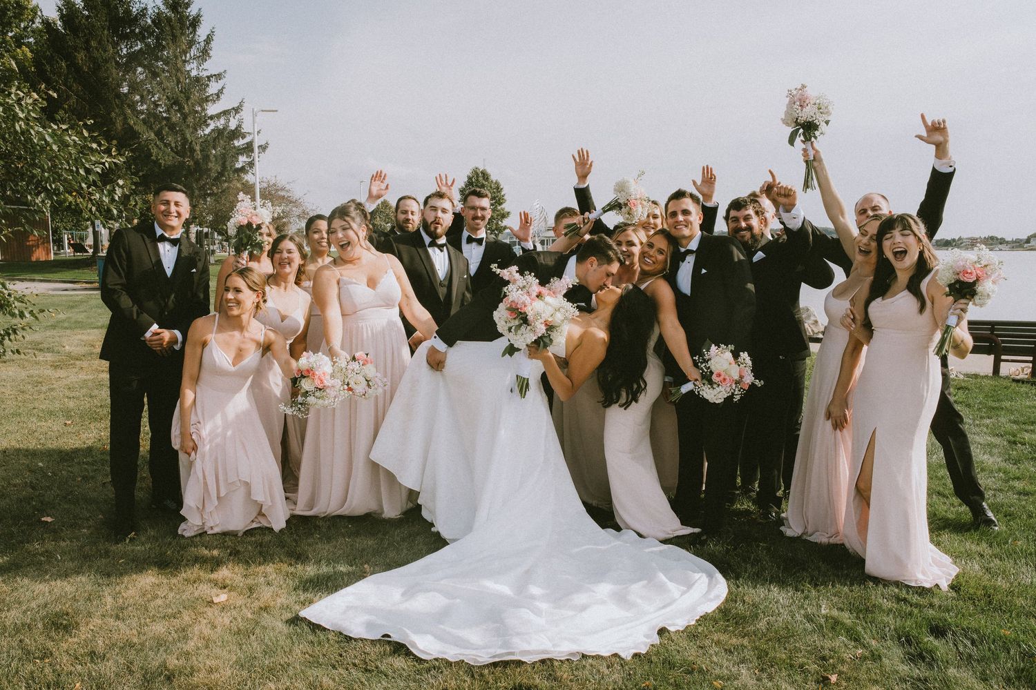 Wedding party in formal attire poses together outdoors on a sunny day with trees and grass in the background.