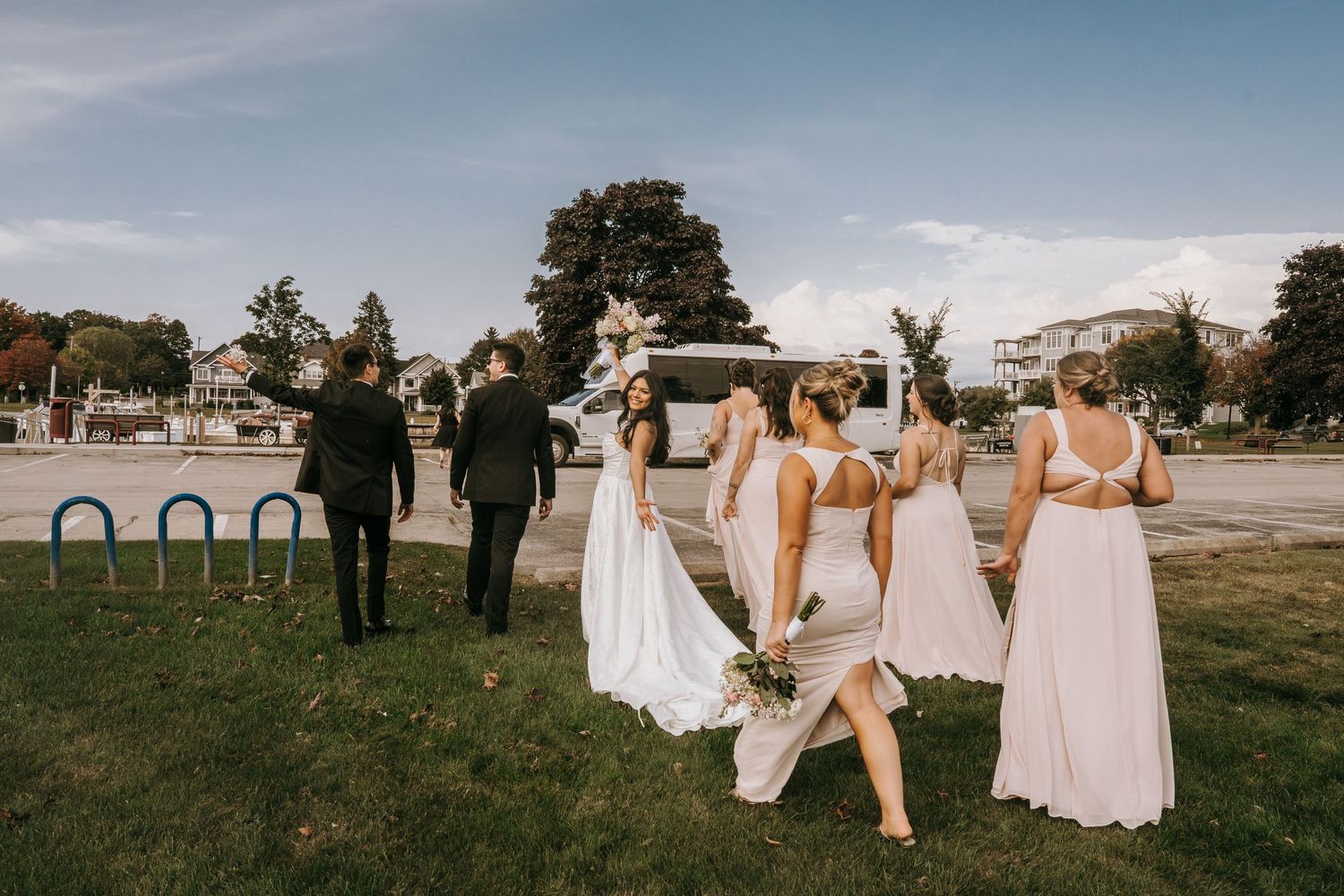 Wedding party walking across green lawn, bride in white gown with bridesmaids and groomsmen heading to venue.