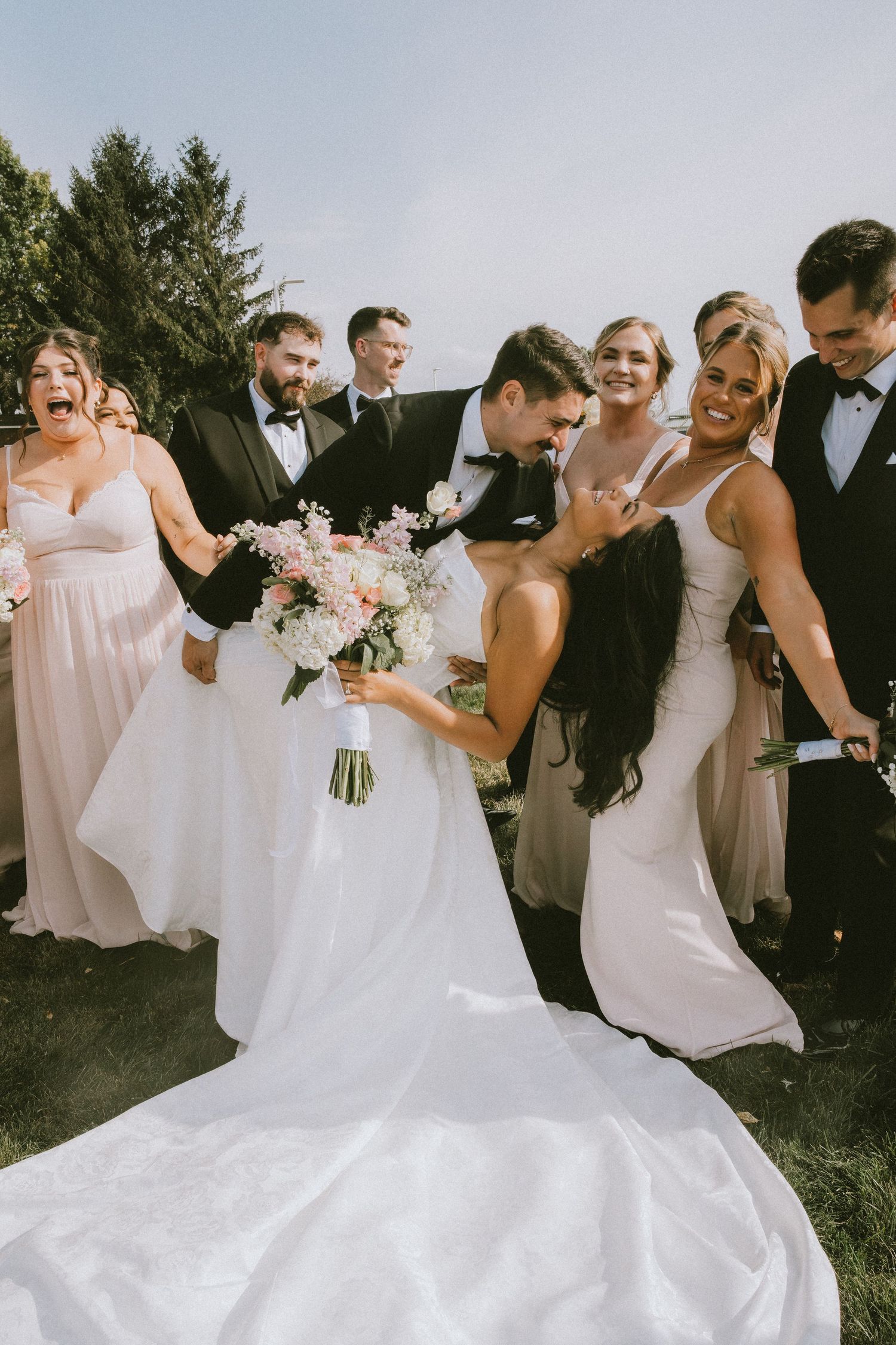 A joyful wedding party in light pink bridesmaid dresses and dark suits celebrates outdoors on a sunny day.