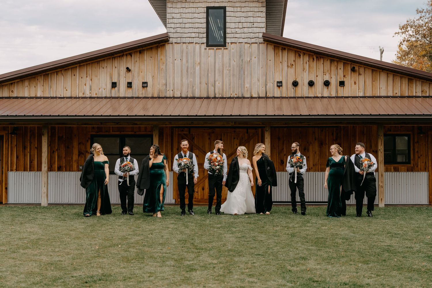 Wedding party in formal attire stands in line formation against rustic wooden barn exterior on grass lawn.