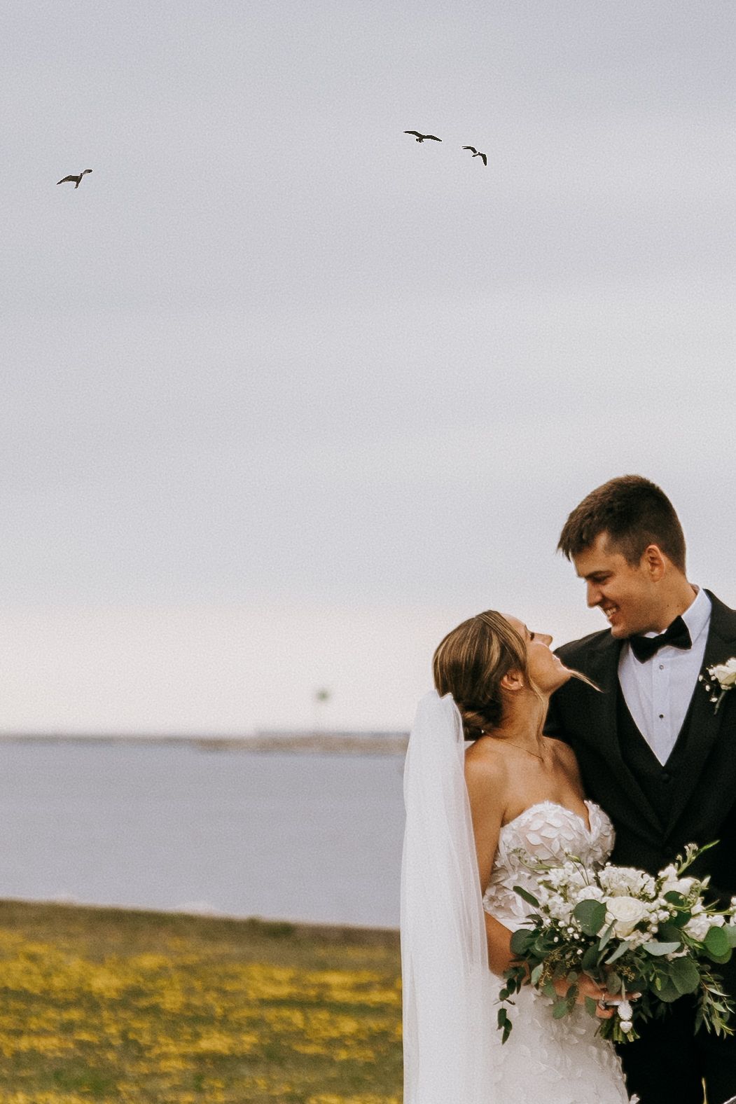 A couple shares a romantic moment by the ocean with birds flying overhead and a bouquet of white flowers.