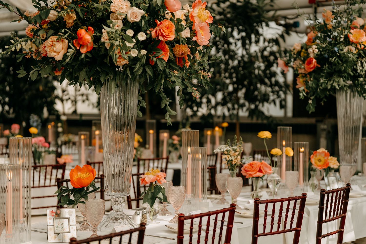 Wedding reception setup with wooden Chiavari chairs and hanging floral arrangements in orange and peach tones.
