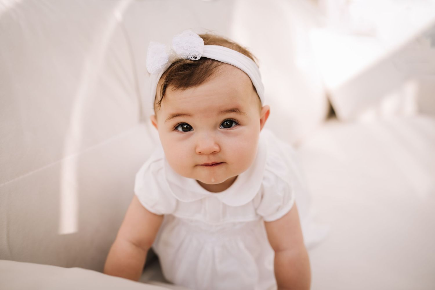 Baby wearing white dress and headband smiles while sitting against light background.