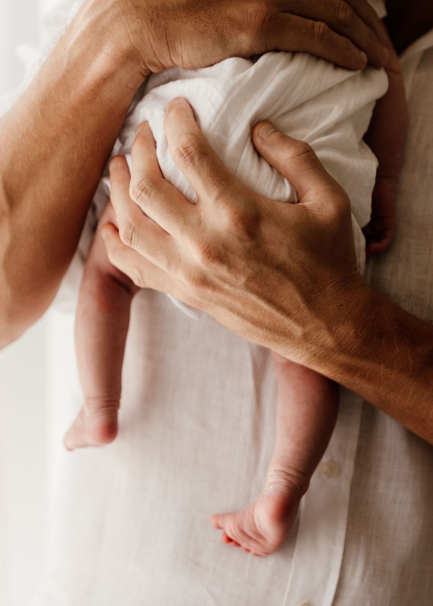 Close up of dad's big hands holding tiny newborn baby against his chest with baby's legs dangling down. soft neutral colours
