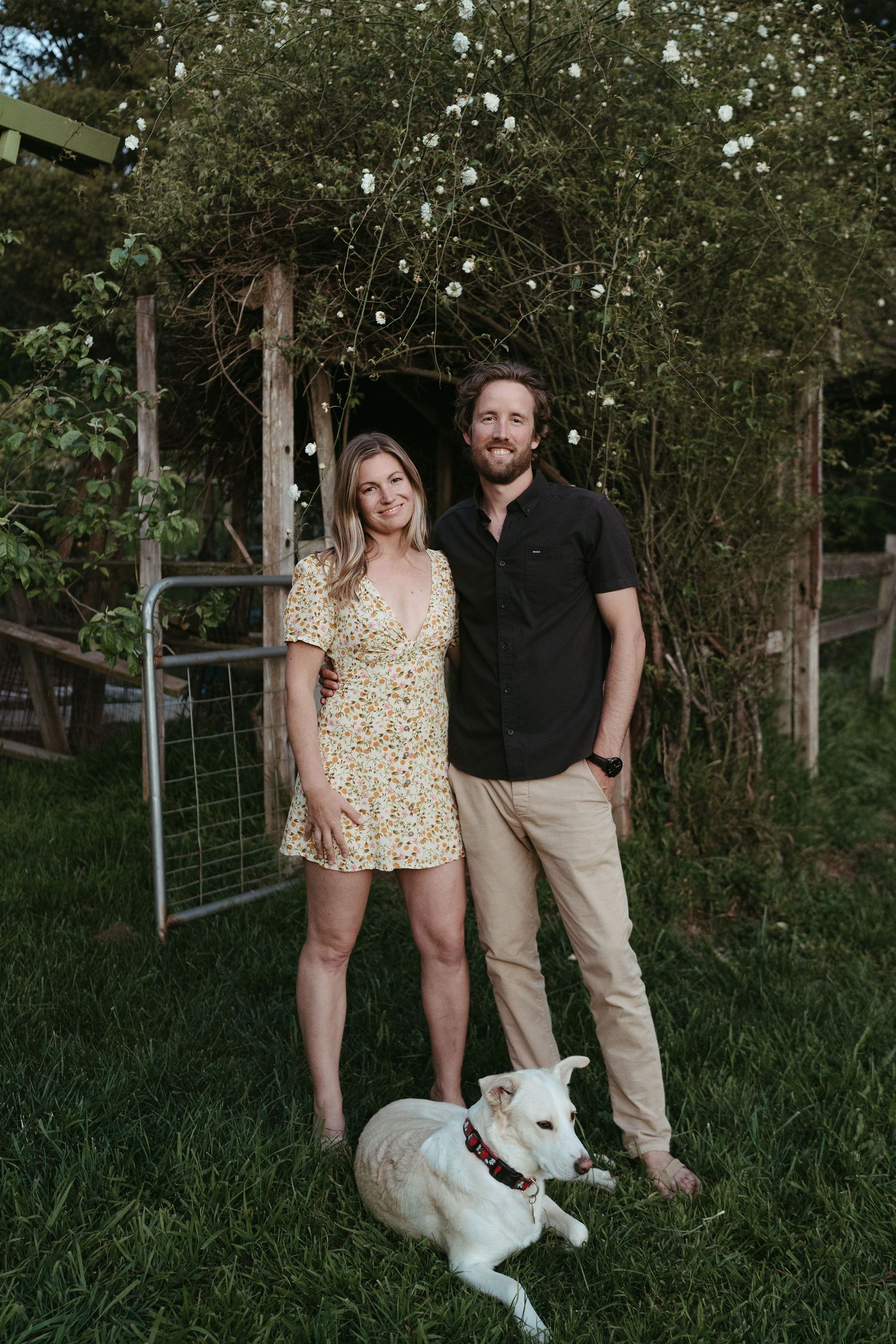 Couple stands with their white dog in front of garden trellis and greenery.