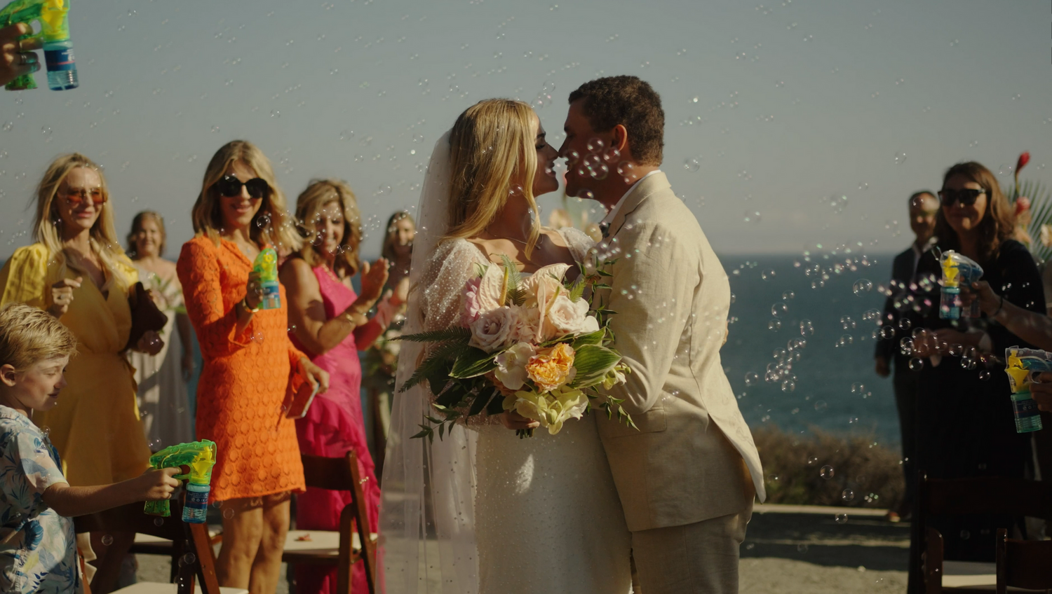 A wedding ceremony on a coastal overlook with guests in bright dresses watching the couple.