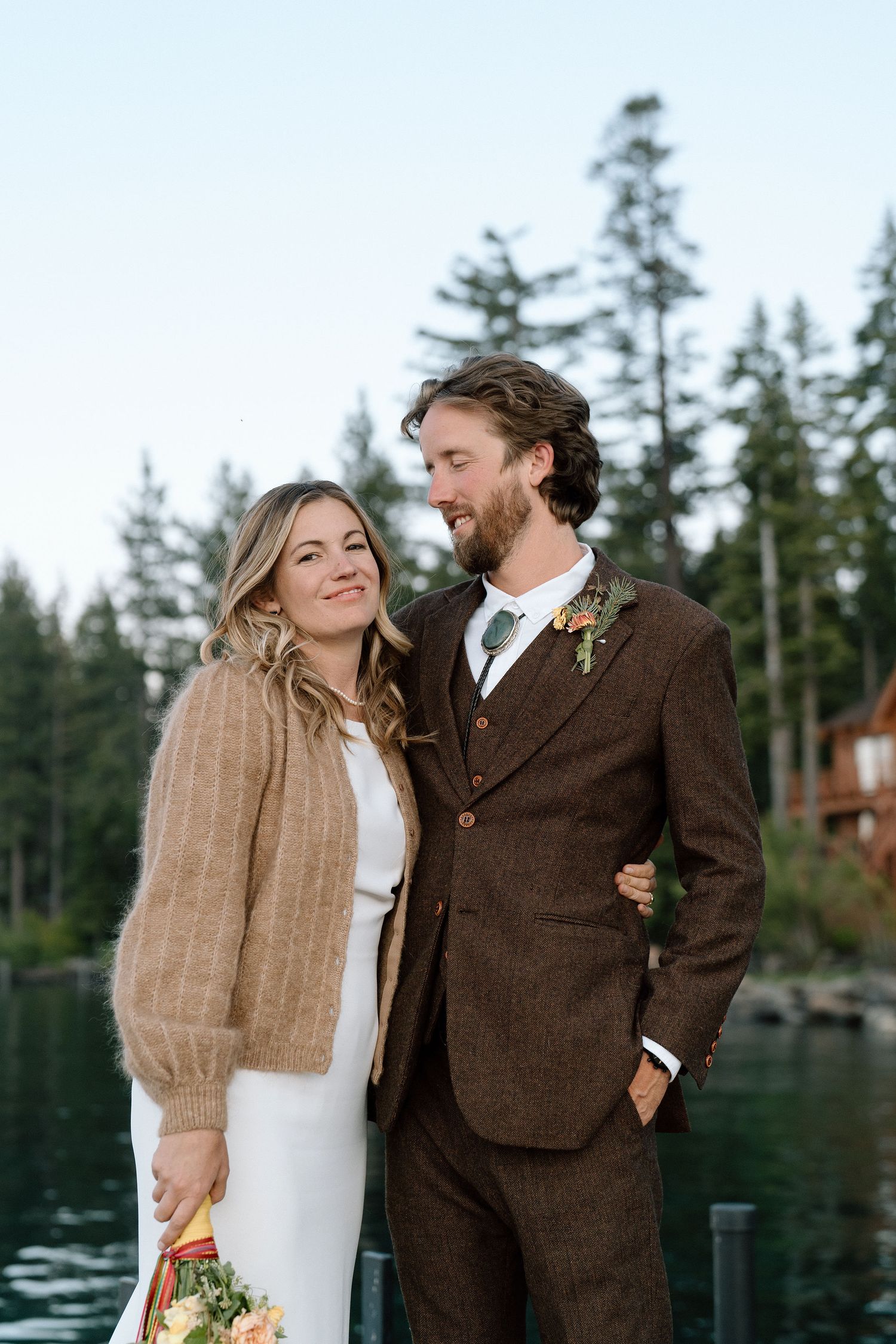 A couple in wedding attire embrace by a lake with evergreen trees in the background.
