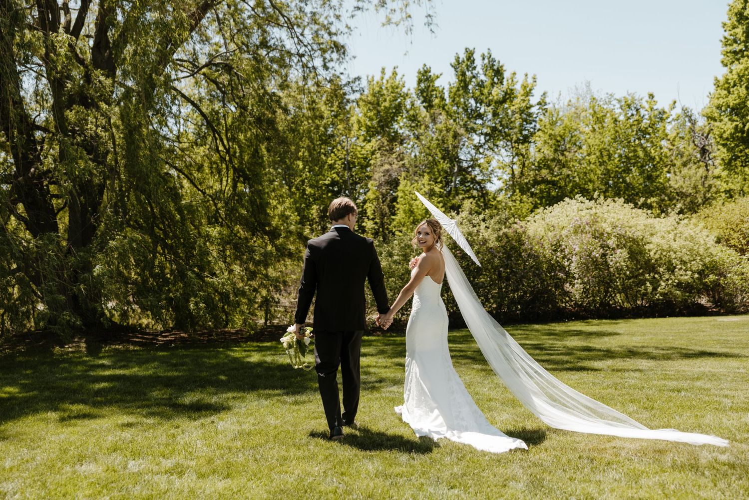 A bride and groom walk hand-in-hand across a lush green lawn surrounded by trees on their wedding day.