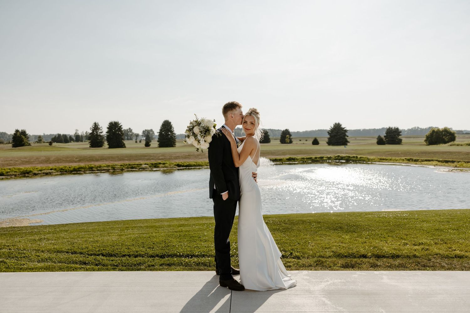 A bride and groom stand together on a grassy field near a body of water on their wedding day. Fort Worth Texas wedding photographer.