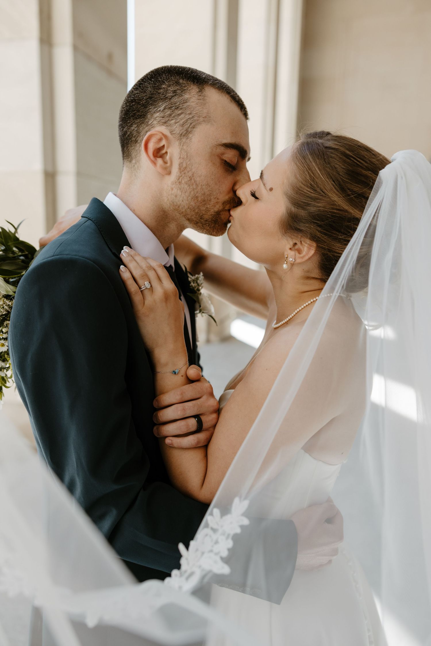 A bride and groom share a romantic kiss, with her white lace veil flowing, as she holds his face tenderly.