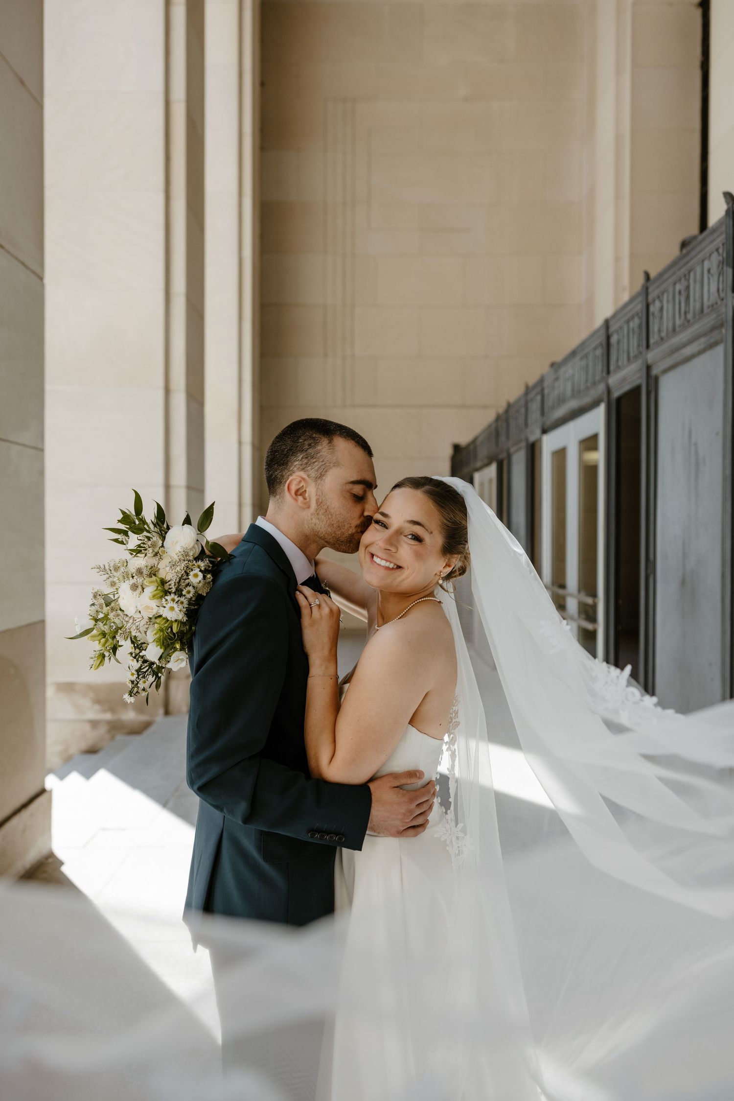 A bride in a flowing veil smiles as her groom kisses her cheek at an elegant columned venue.