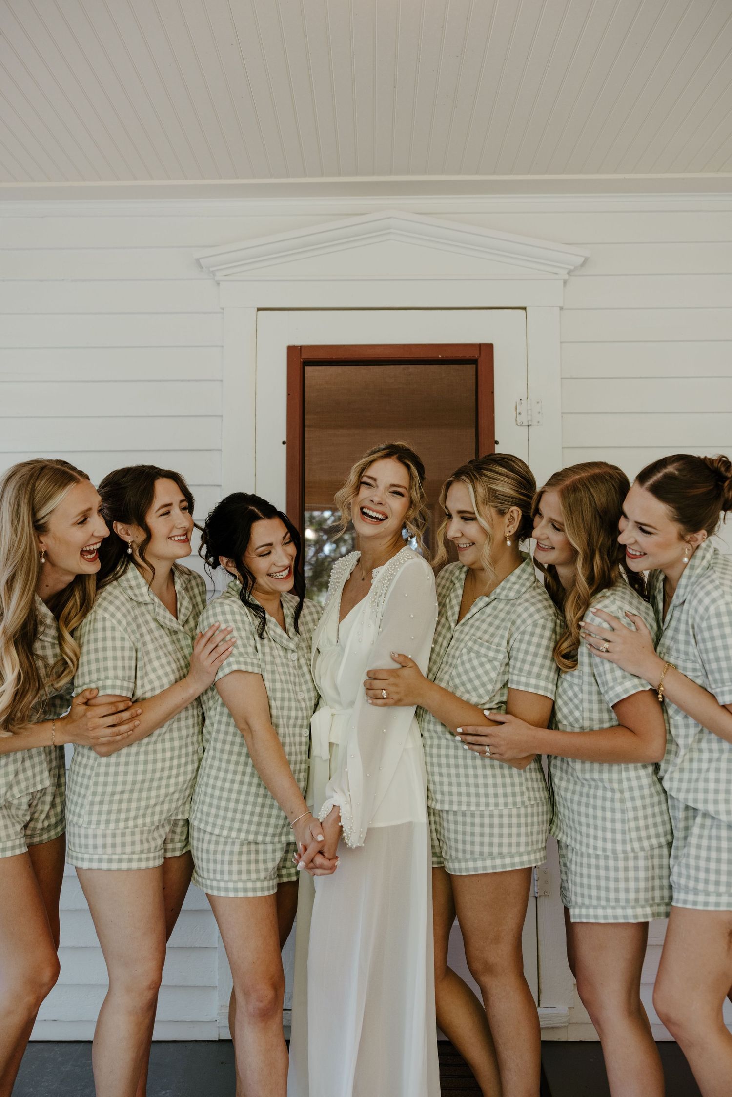 Bride in white robe laughing with bridesmaids in matching green gingham pajama sets during wedding morning prep.