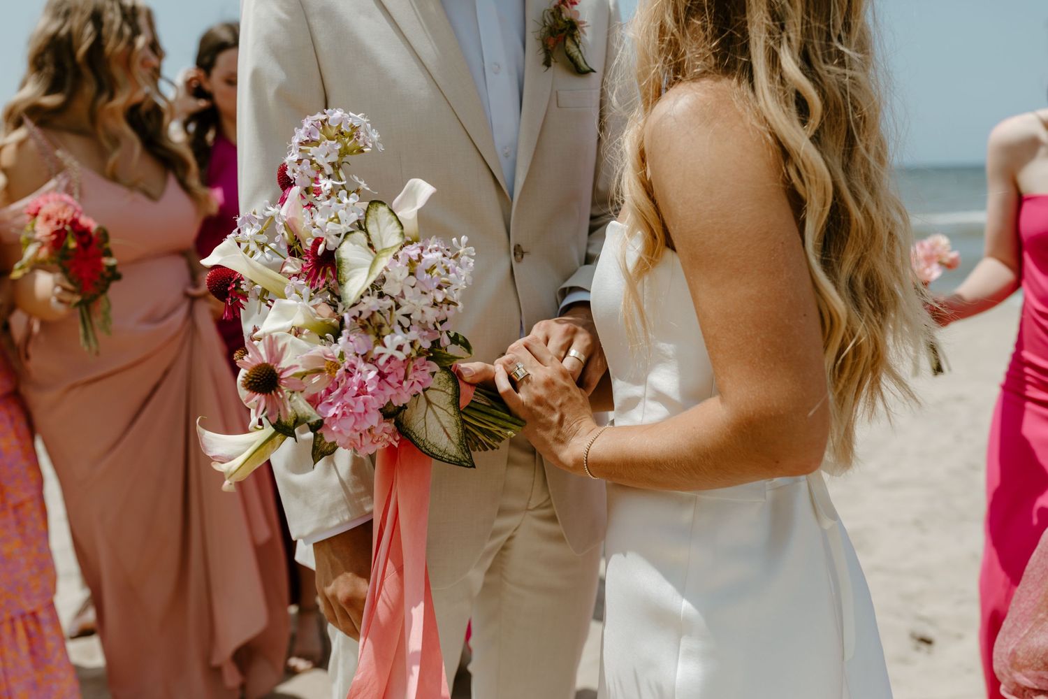 A bride and groom stand together, holding a bouquet of flowers, surrounded by their wedding party in colorful dresses.