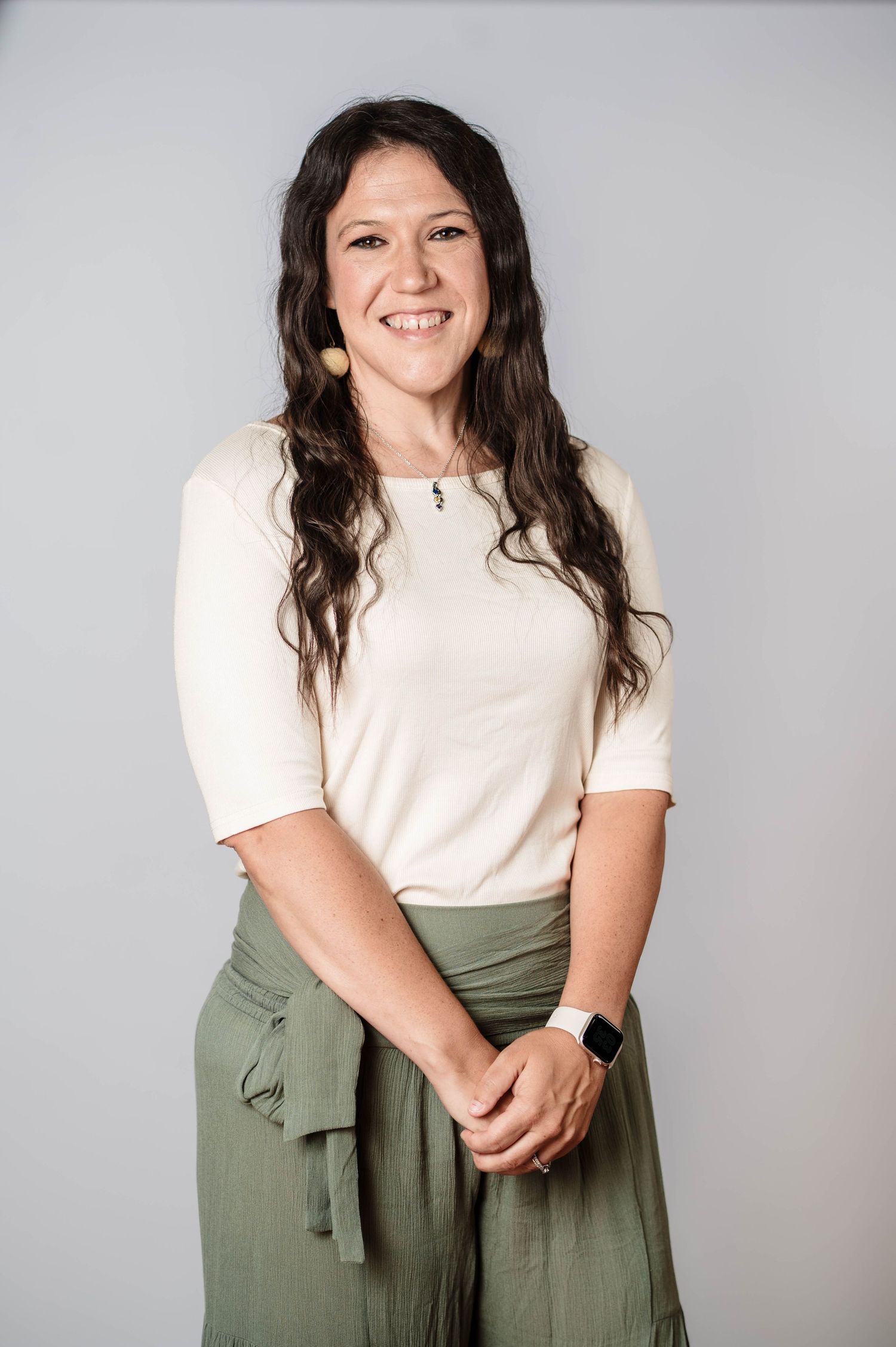 Smiling woman with dark wavy hair wearing a cream top and olive green skirt, posing against a gray background.