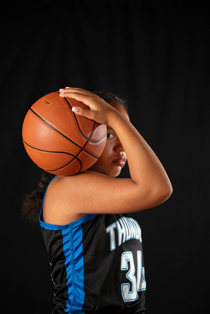 Young girl in Thunder #34 jersey holds basketball in shooting position against black background.