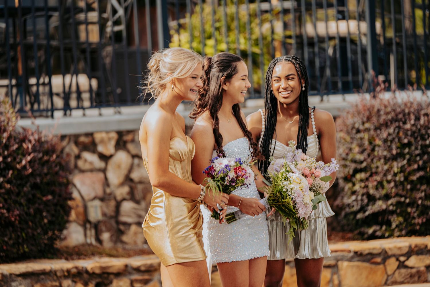 Three bridesmaids in elegant dresses holding bouquets, smiling outdoors near a stone wall.