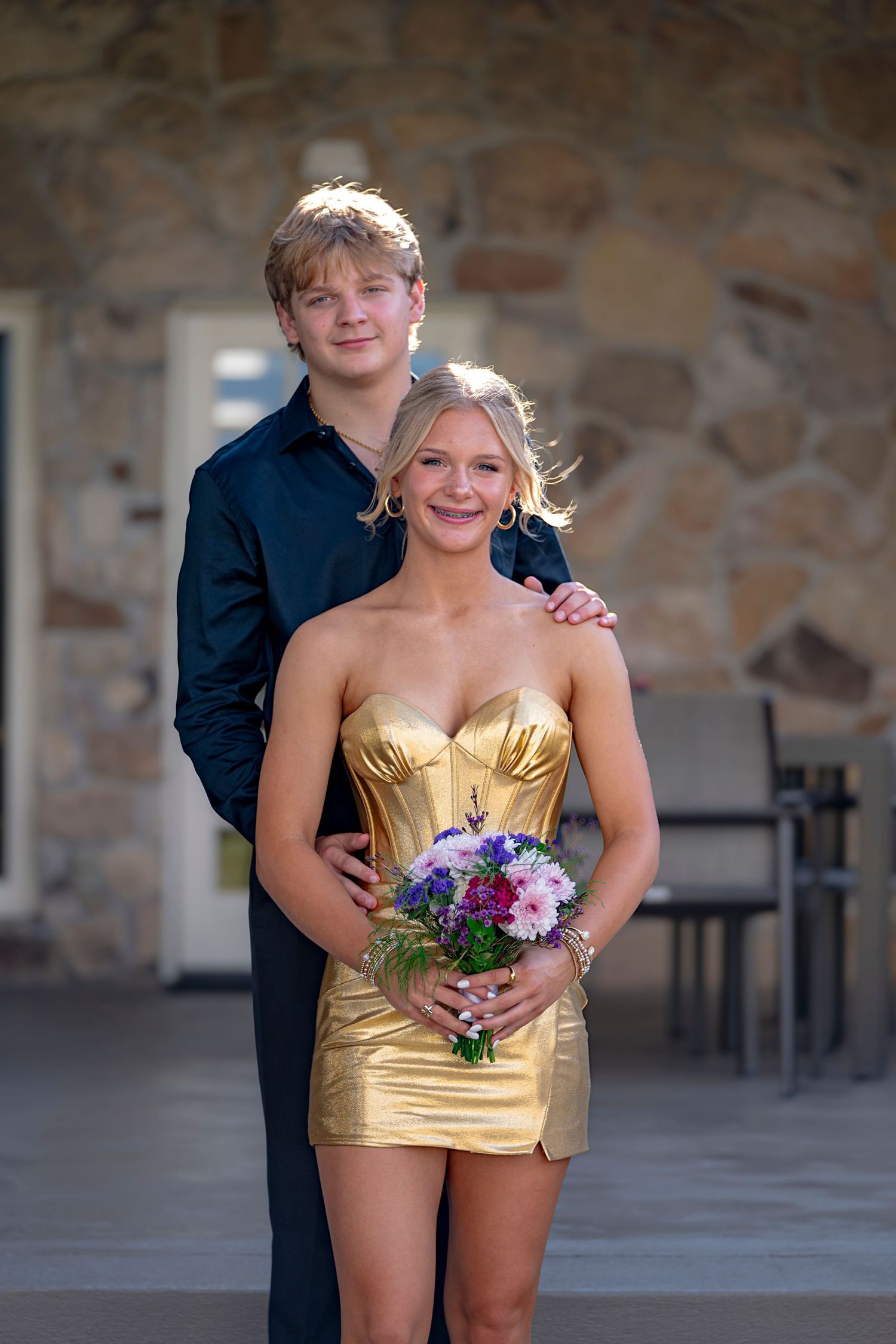 Teen couple dressed for prom, girl in gold mini dress holding bouquet, boy in black shirt, posing outdoors.