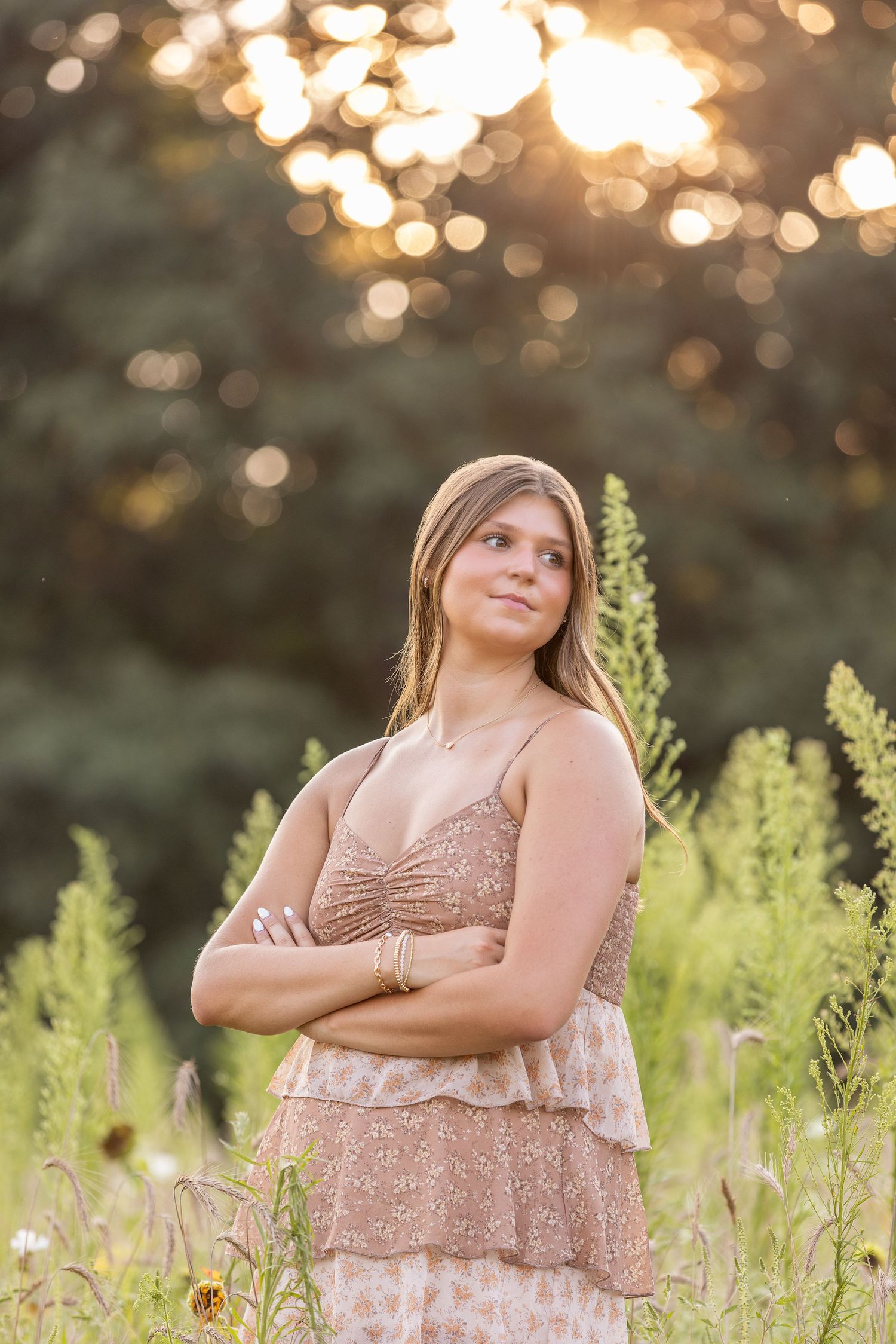 Young woman in floral dress standing in a sunlit wildflower field with golden bokeh background at sunset.