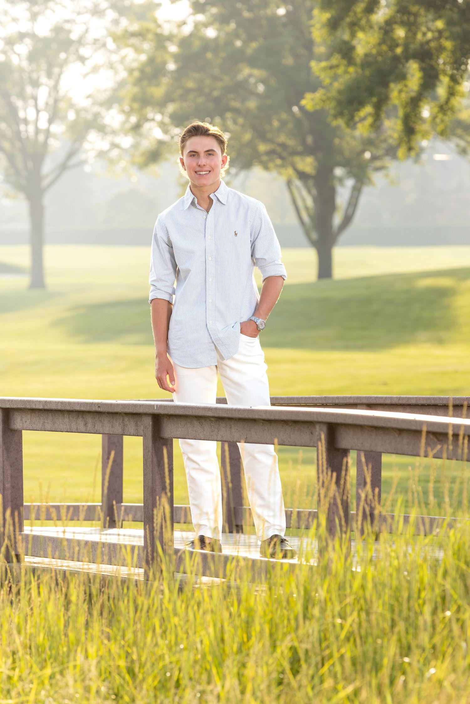 Young man in white pants and striped shirt smiling on a wooden bridge surrounded by lush green grass and trees.