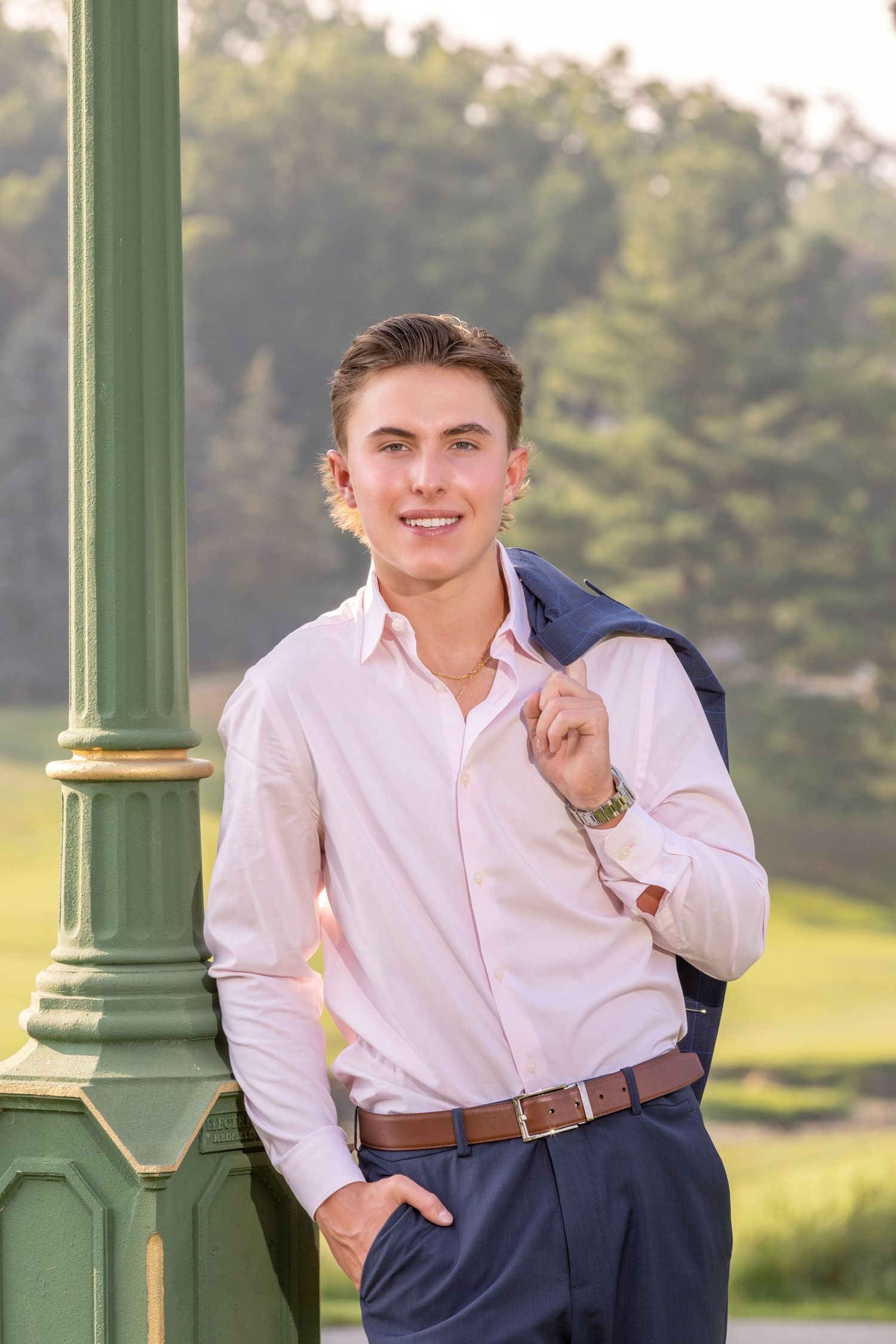 Young man in pink shirt and navy suit jacket smiling outdoors near green lamppost with lush greenery background.