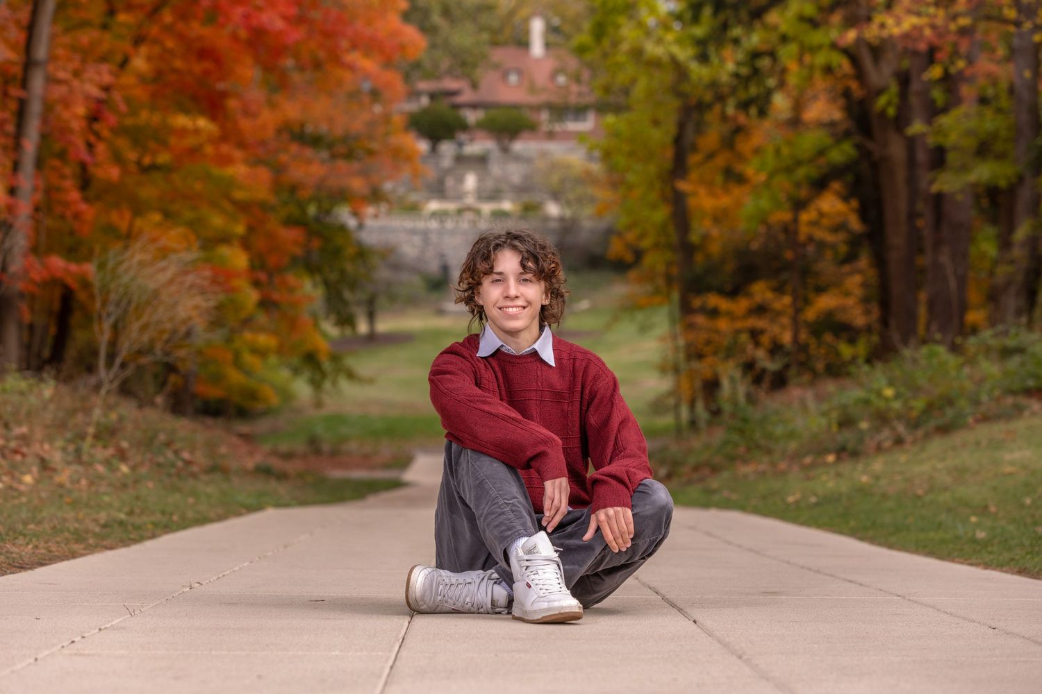 Teen sitting cross-legged on a path surrounded by vibrant fall foliage trees, wearing a red sweater and white sneakers.