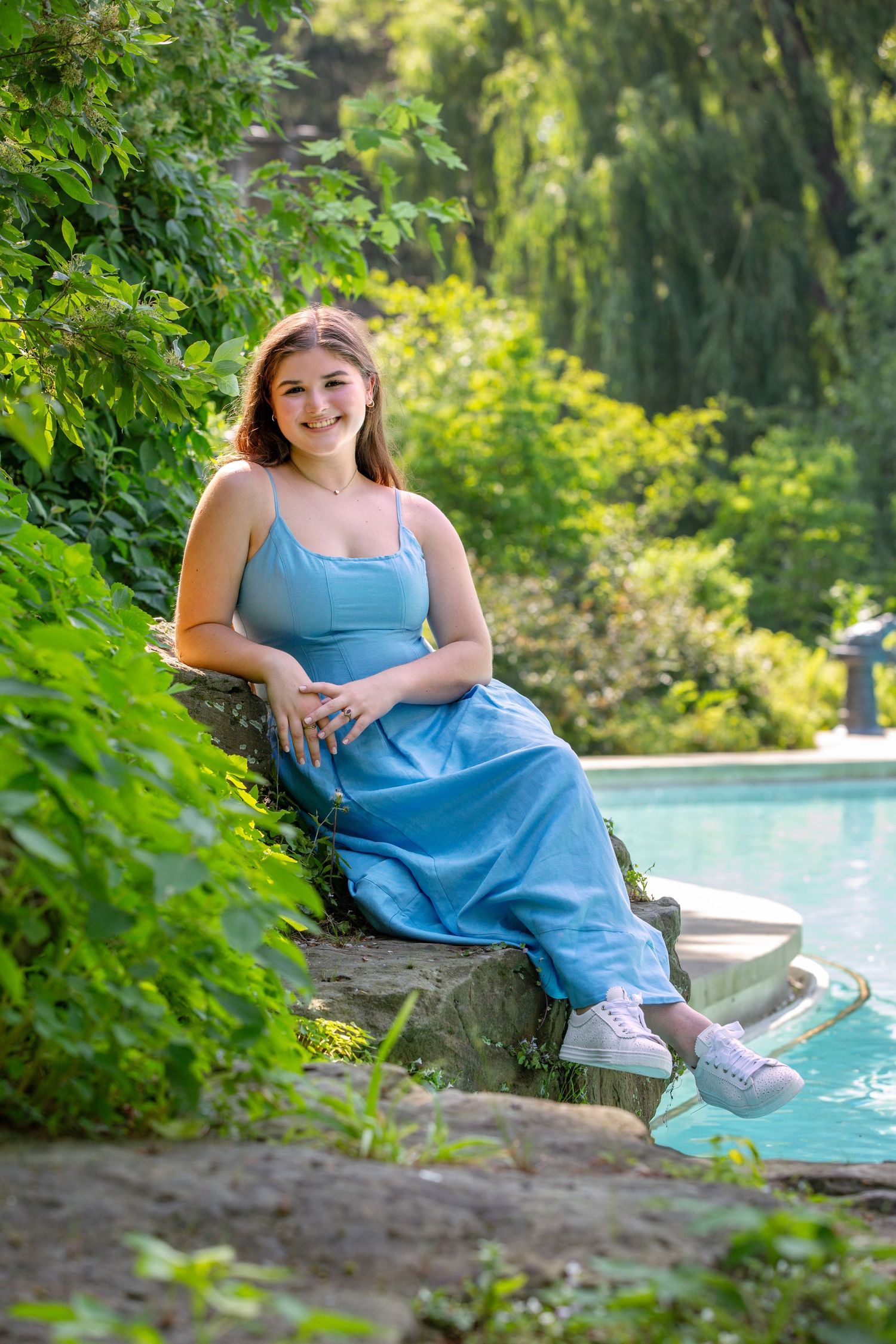 Someone in a blue outfit sits on rocky landscaping beside a pool surrounded by lush summer greenery and willow trees.