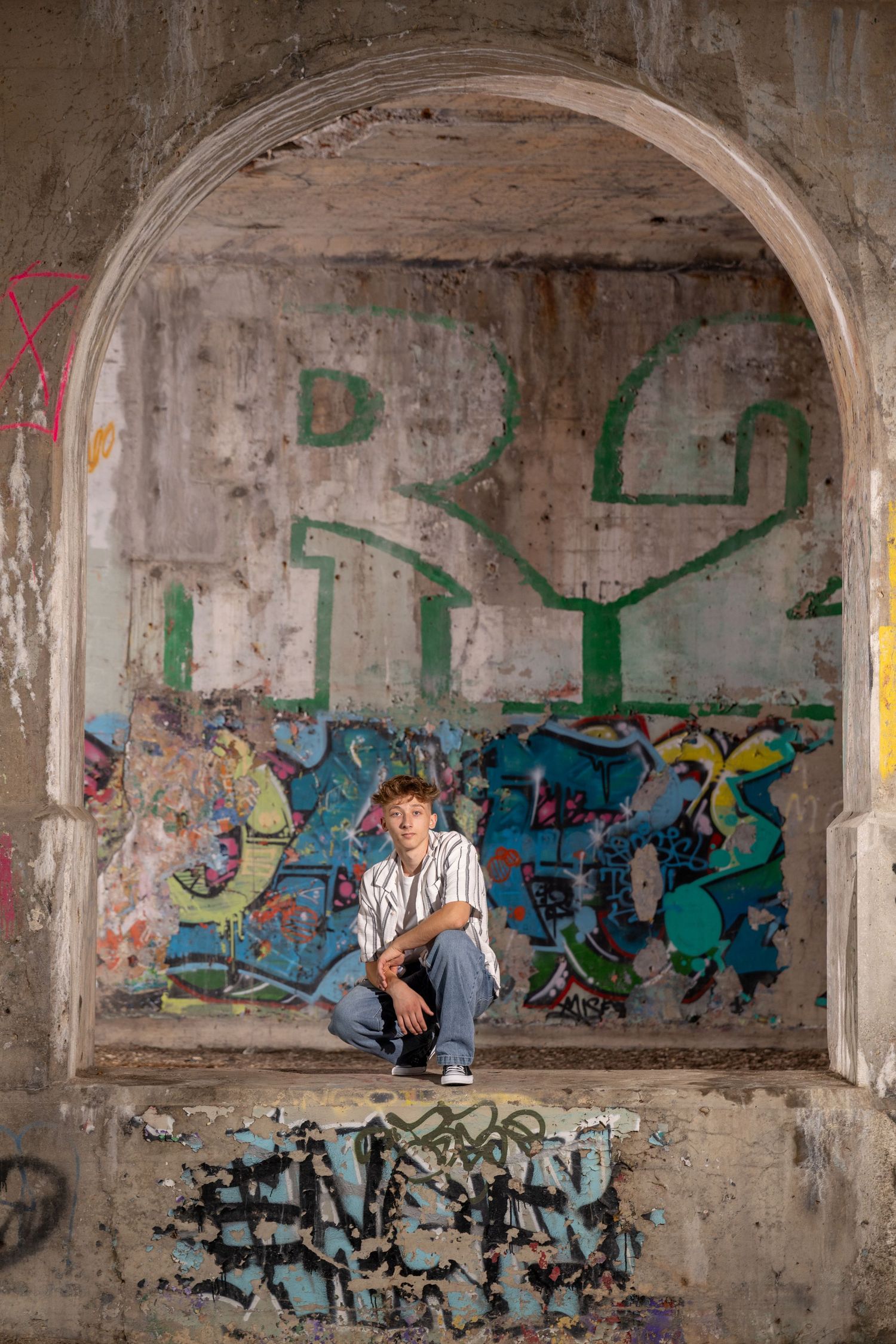 Young man crouching in graffiti-covered arched alcove of an abandoned urban building.