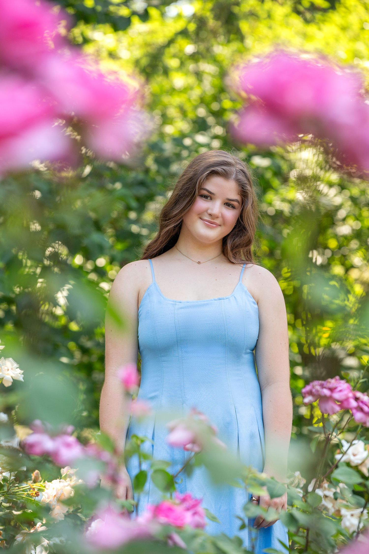Young woman in light blue sundress smiling among pink roses in a lush green garden on a sunny day.