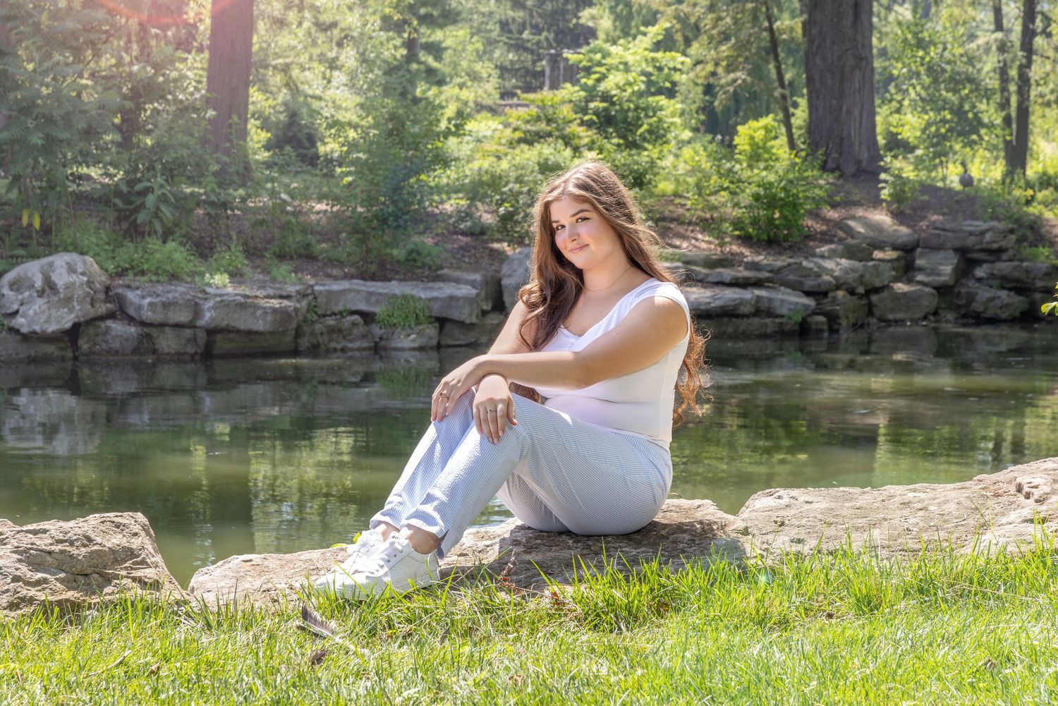 Young woman in white top and jeans sitting on rocks by a peaceful pond in a lush green park.