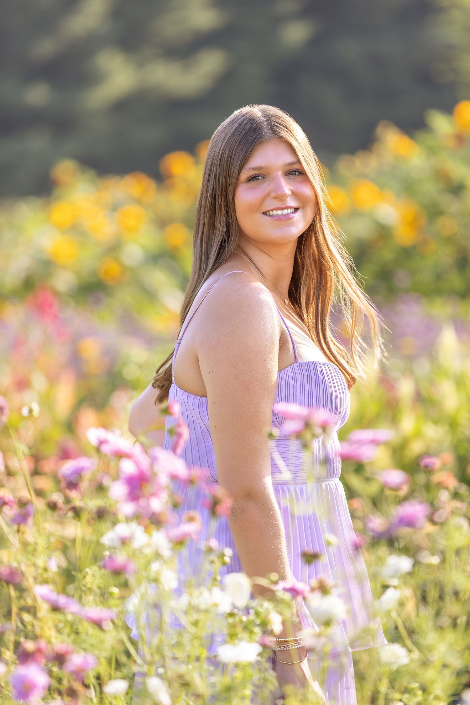 Smiling young woman in lavender sundress standing among colorful wildflowers in a sunny garden.