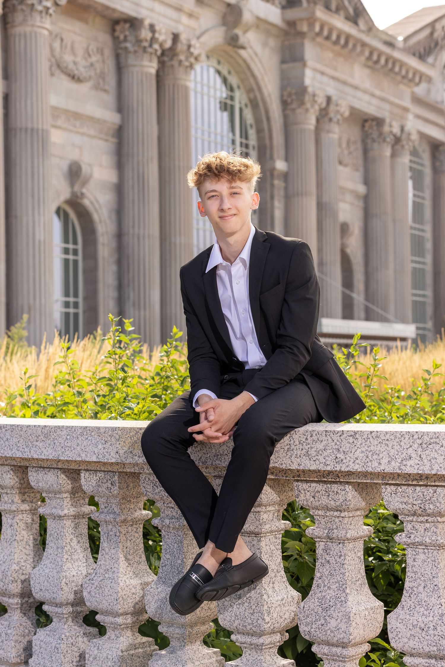 Young man in black suit sitting on stone balustrade outside classical building with columns.