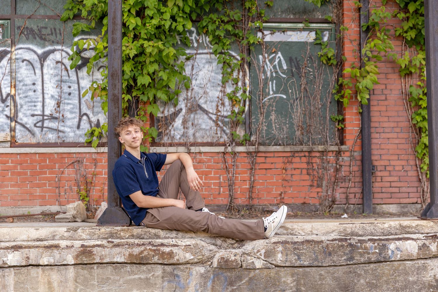 Young man sitting on a concrete ledge against a brick wall with ivy and graffiti in an urban setting.