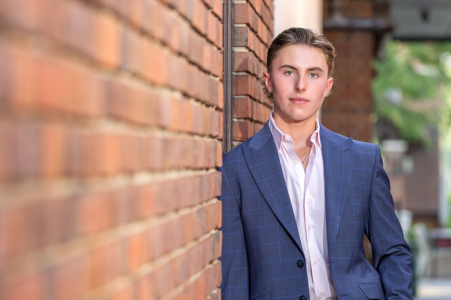 Young man in blue blazer leaning against a brick wall, posing for a professional senior portrait photo.