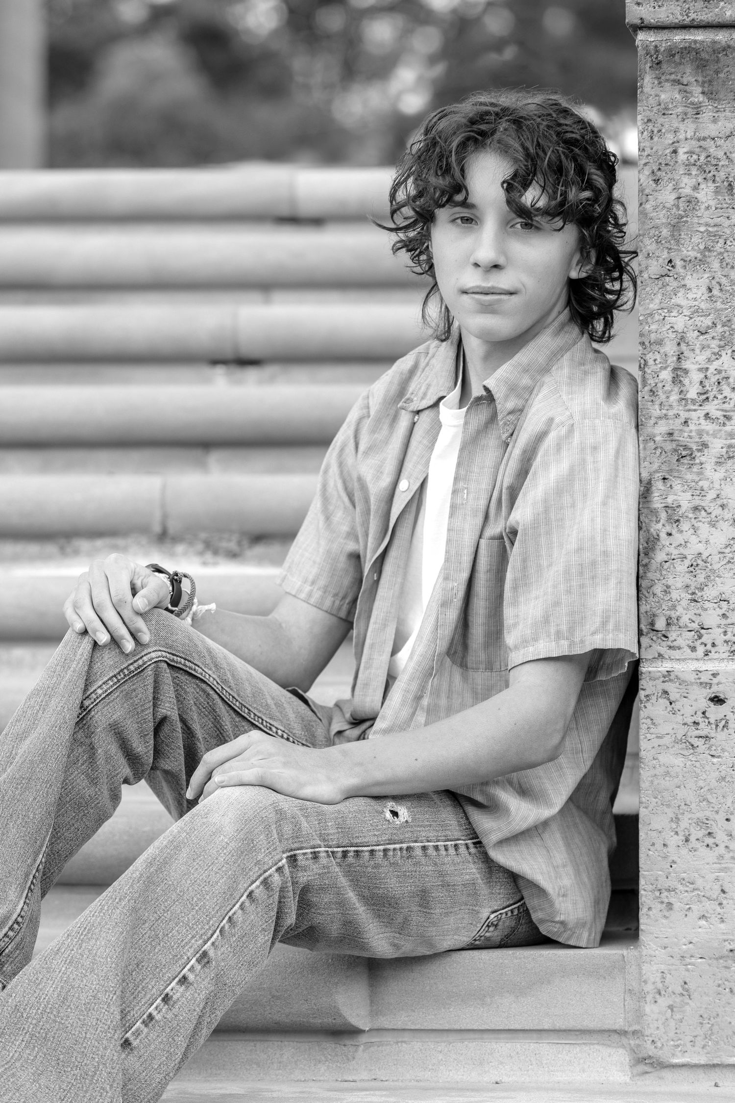 Black and white portrait of a teenage boy with curly hair sitting on steps, wearing a striped shirt and jeans.