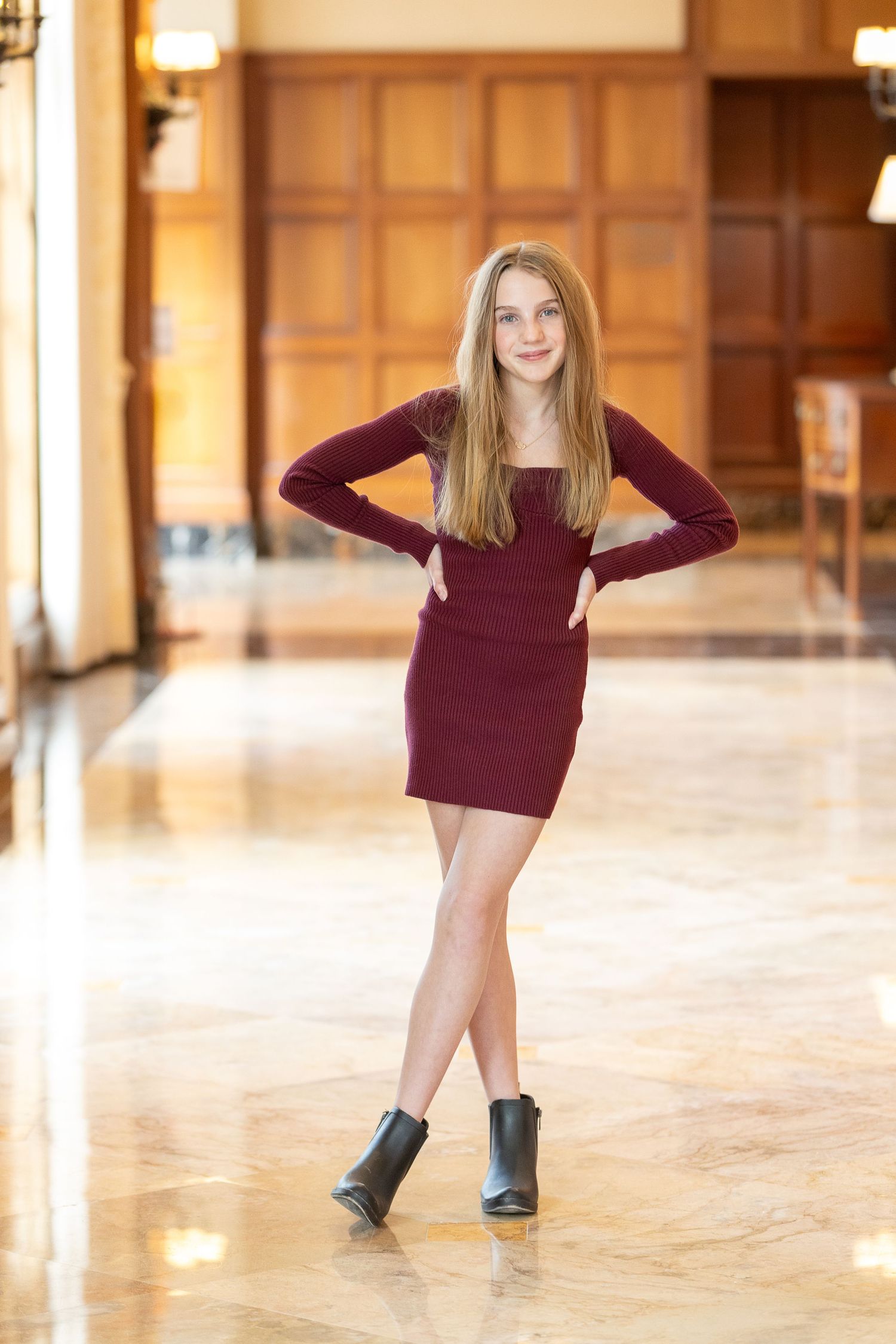 A person in a burgundy long-sleeved dress and ankle boots poses confidently on a polished floor in an elegant hallway.