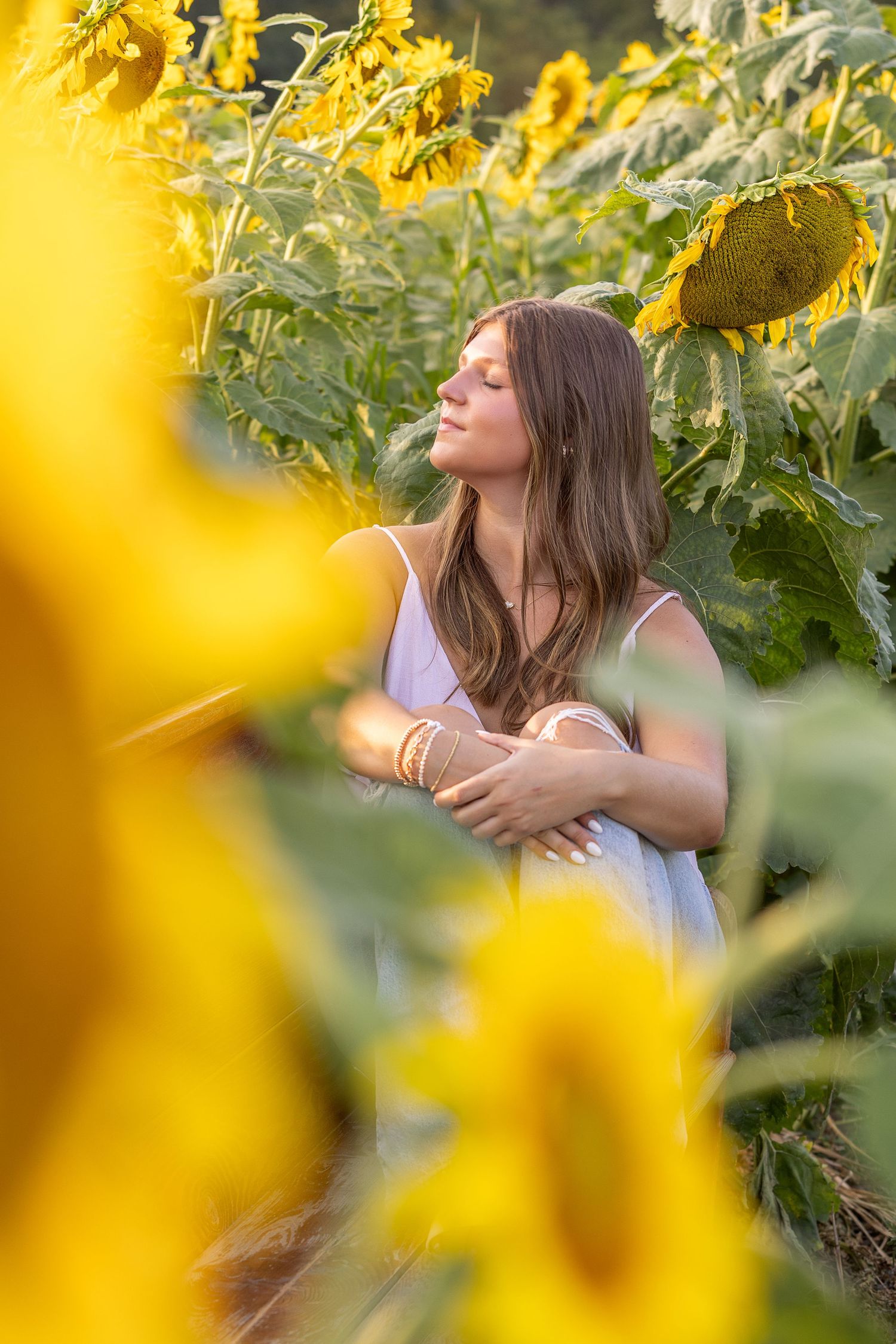 Young woman with eyes closed embracing herself peacefully among tall sunflowers in golden light.