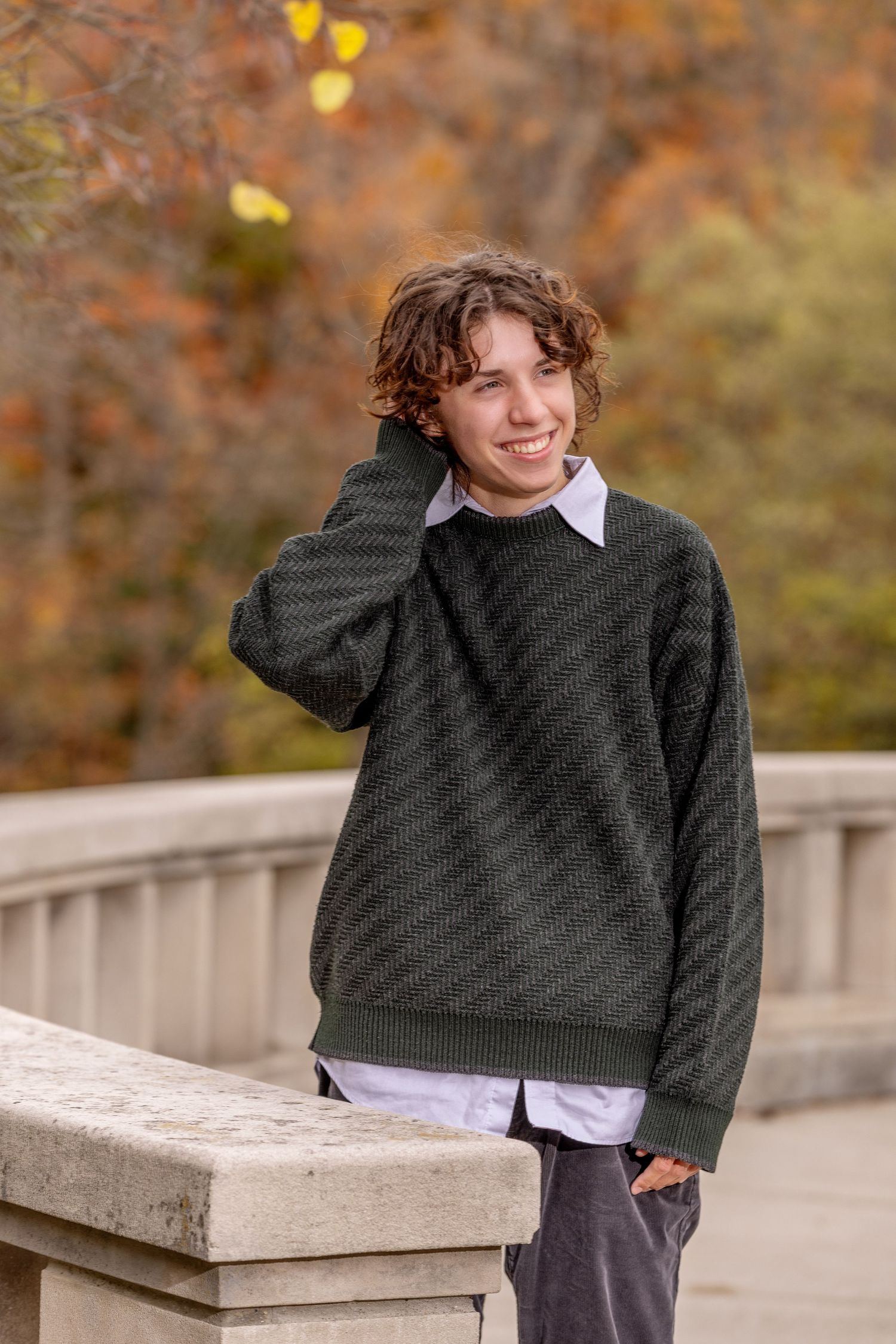 Smiling teenage boy in dark green sweater over white shirt poses on stone bridge amid autumn foliage.