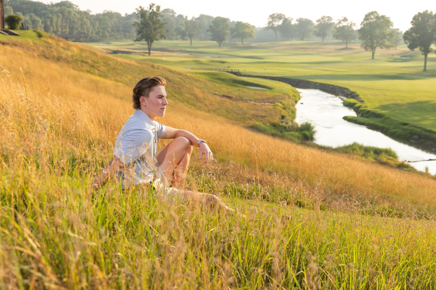 Young woman sitting in tall golden grass beside a winding stream on a lush golf course at sunset.