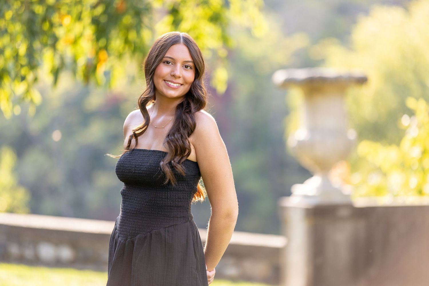 Young woman in black strapless dress smiling outdoors in lush green garden setting with stone urn.